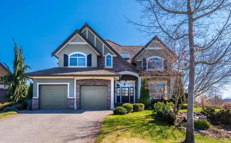 Large two-story house with stone facade, two-car garage, and landscaped lawn under a clear blue sky.