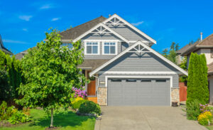 Exterior of a suburban two-story house with grey siding, double-garage, green lawn, and sunny blue sky.